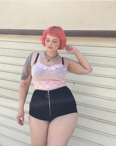 a woman with red hair wearing black shorts and a pink top is leaning against a wall