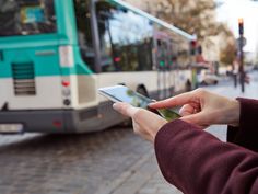 a person holding a cell phone in front of a bus on a street with people walking by