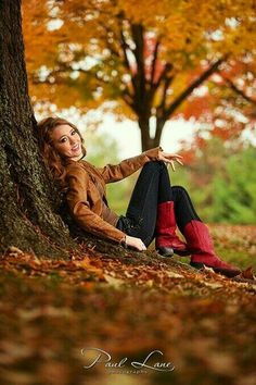 a beautiful woman sitting on the ground next to a tree in an autumn park with leaves all around her