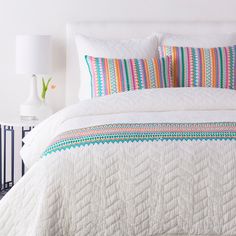 a white bed with colorful pillows and quilts on it's headboard next to a night stand