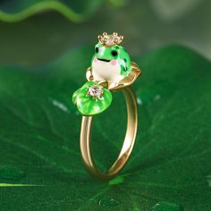 This whimsical piece captures the essence of nature's beauty and adds a touch of regal charm to your ensemble. Made from sterling silver and hand painted with enamel, this enchanting ring features an adorable frog wearing a crown, poised gracefully amidst a lotus leaf. The intricate flawless enamel work coupled with white stones, make this an eye-catching piece. This exquisite combination of elements evokes a sense of tranquility and harmony. It will surely become a conversation starter and a cherished addition to your jewelry collection.Carat Weight: 0.545 ctStone Size: 2.5,3.5 mmStone Type: Jeulia® StoneNumber of Stones: 3 Stone Shape: RoundStone Color: Diamond WhiteWeight: 5.8 gWidth: 2.2 mmHeight: 10.4 mmThickness: 1.3 mmMaterial: 925 SilverPlating Color: Yellow Gold Wearing A Crown, Frog Jewelry, 9th Anniversary, Frog Prince, White Stones, Lotus Leaves, Lotus Leaf, Anniversary Sale, White Stone