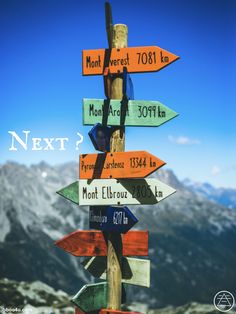 a wooden sign pointing in different directions with the words next? written on it and mountains in the background