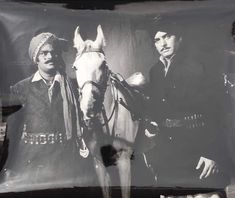 two men standing next to each other with a horse in the foreground and an old photo on the wall behind them