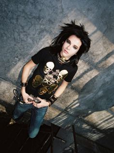 a woman with black hair standing in front of a concrete wall and holding her hand on her hip