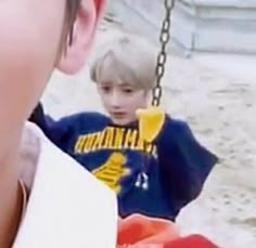 a young boy sitting on a swing in the sand with another child standing behind him