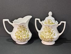 two white vases with gold designs on them sitting next to each other in front of a gray background