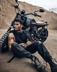 a man sitting on the ground next to a motorcycle with his legs crossed and wearing boots