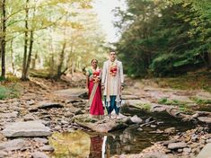 a man and woman standing on rocks in the middle of a stream with trees behind them