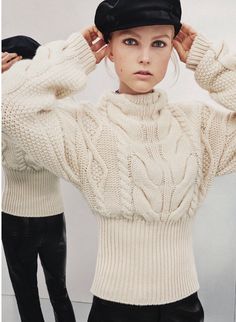 a woman in white sweater and black hat holding her hands on her head while standing next to another person