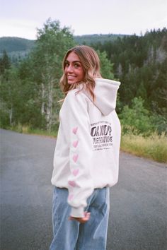 photo shows a brunette teenage girl walking on a paved road in the mountains on a cloudy day. she is wearing blue jeans and a gray hoodie that has pink hearts embroidered on the sleeves. the girl is walking away from the camera but is looking back at the camera and smiling. there are green bushes and trees in the background as well as hills. photo was taken on film Heart On My Sleeve Dandy Hoodie, Heart On My Sleeve, Bday Wishlist, Cute Jumpers