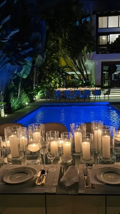 a table set with candles and place settings next to a swimming pool at night time