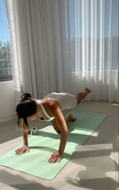 a woman in white shirt and black pants doing yoga on green mat with window behind her