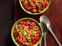 two green bowls filled with chili and cheese on top of a wooden table next to spoons