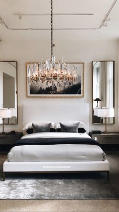 a white bed sitting under a chandelier next to two large mirrors on the wall