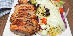 a white plate topped with meat and vegetables next to a bowl of dressing on top of a wooden table