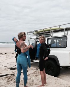 two men in wetsuits standing next to a white van on the beach