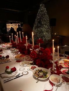 a table set for christmas dinner with candles and plates full of food in front of the tree