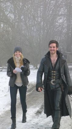 a man and woman dressed in medieval clothing walking down a snowy path with trees in the background