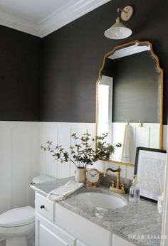 a white bathroom with black walls and gold trim on the mirror above the sink is decorated with greenery