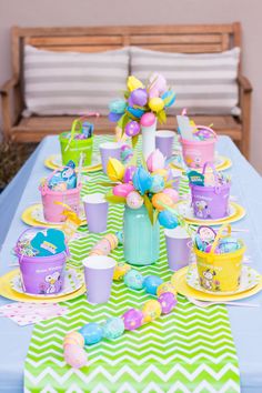 a table topped with lots of cups and plates filled with colorful candy eggs on top of a green and white striped table cloth