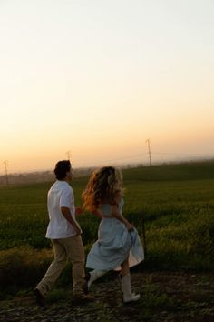 a man and woman walking through a field at sunset