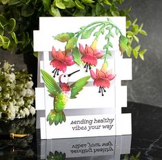 a card with flowers and a hummingbird on it, sitting next to a plant