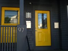 a yellow door is on the side of a gray house with black siding and windows