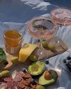 two glasses of wine, cheese and fruit on a table with other food items in the background