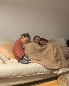 three people laying on a couch covered in blankets and pillows, all looking at the camera