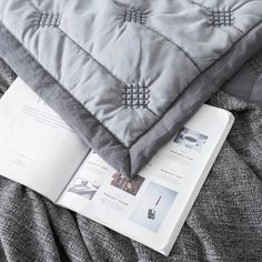 an open book on top of a bed next to a gray quilted pillow and blanket