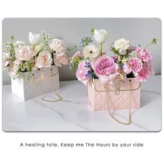 two pink and white flowers are in small boxes on a marble countertop, one is holding a gold chain