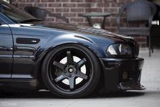 the front end of a black car parked in front of a brick building with tables and chairs behind it