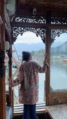 a woman standing in front of a window looking out at the water and mountains behind her