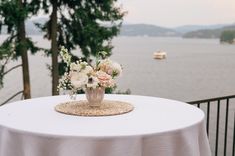 a vase with flowers sitting on top of a white table cloth covered table next to the water