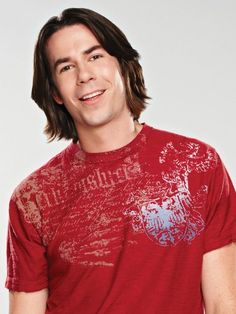 a man with long hair wearing a red t - shirt and smiling at the camera