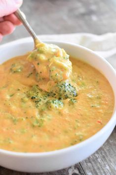 a bowl of broccoli cheese soup with a spoon in it and someone's hand holding a ladle