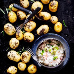 a bowl of potato salad next to some potatoes on a black surface with utensils