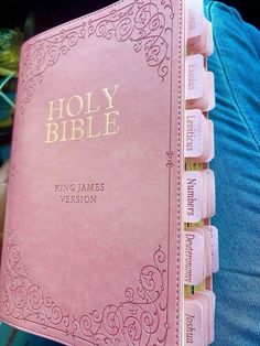 a pink bible with gold lettering is held by someone's hands in blue jeans