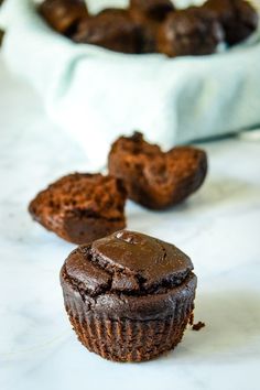 two chocolate muffins sitting next to each other on a white counter top with more muffins in the background