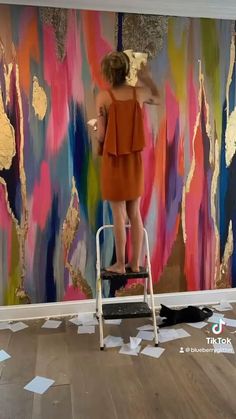 a woman standing on a stepladder painting a wall with gold leafy paint