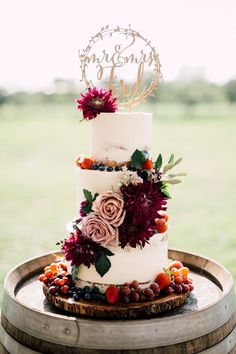 a three tiered wedding cake with flowers and berries on top sits on a barrel