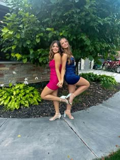 two young women posing for a photo in front of some trees and bushes, one is holding the other's leg