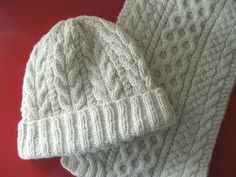 two white knitted hats sitting next to each other on a red surface, one with a cabled design