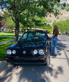 a woman standing next to a parked car on the side of a road near trees