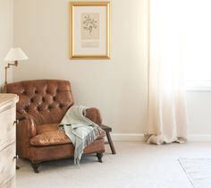 a brown leather chair sitting in front of a window next to a dresser and lamp