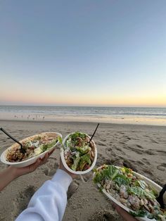 two people holding plates of food on the beach at sunset, with ocean in background