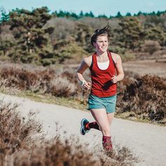 a woman running down a dirt road next to tall grass and bushes on a sunny day