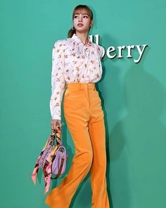 a woman standing in front of a green wall holding a purse and wearing yellow pants