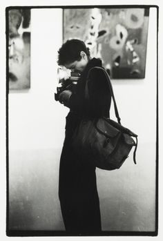 black and white photograph of a man holding a camera in front of paintings on the wall