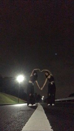 two girls standing on the side of a road at night with their arms in the shape of a heart